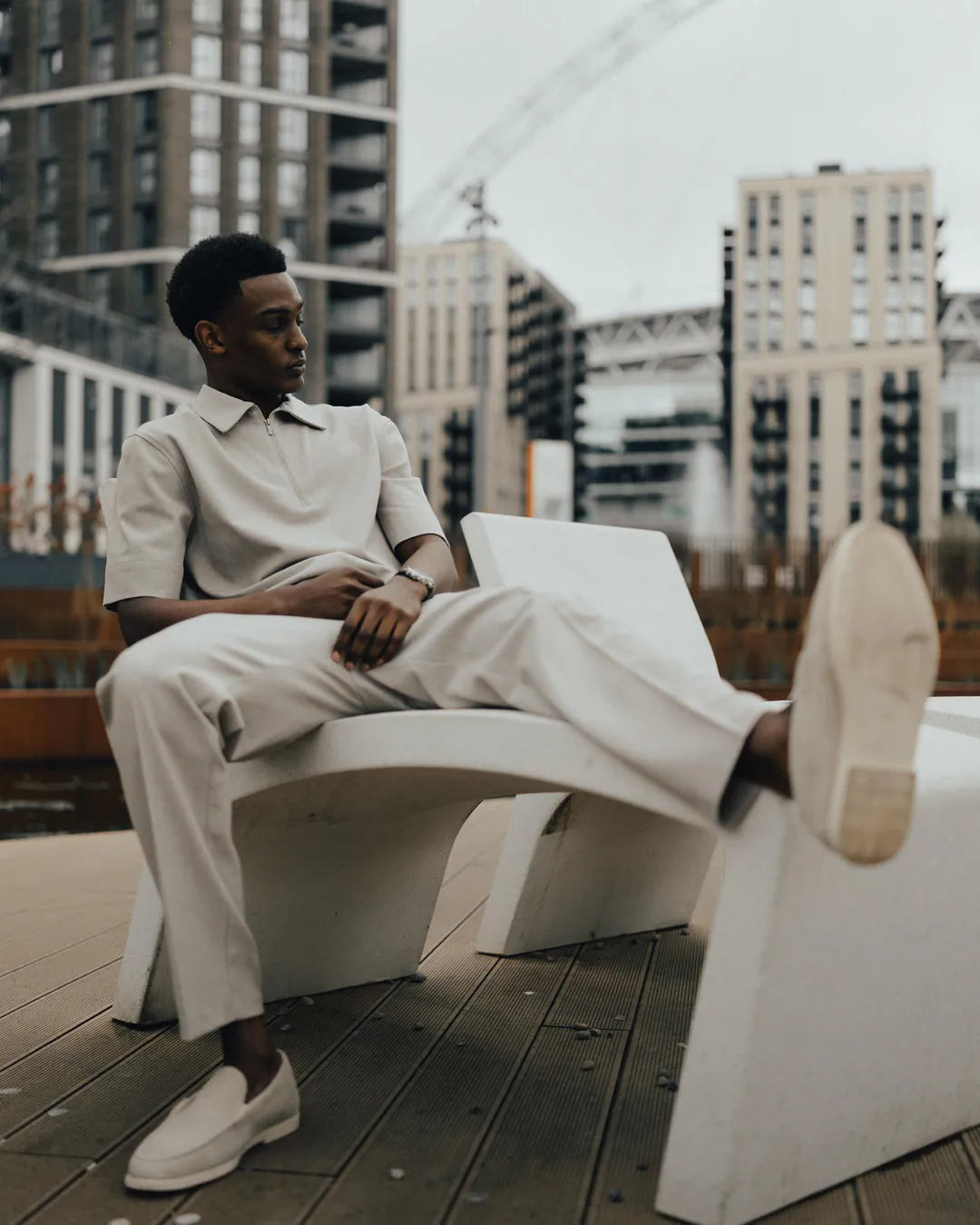 Man in cream shirt and trousers reclining on a white bench with one foot raised, high-rise towers behind