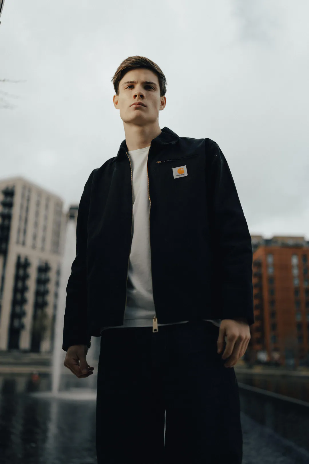 Man in a black workwear jacket and dark trousers standing before a fountain and red-brick flats