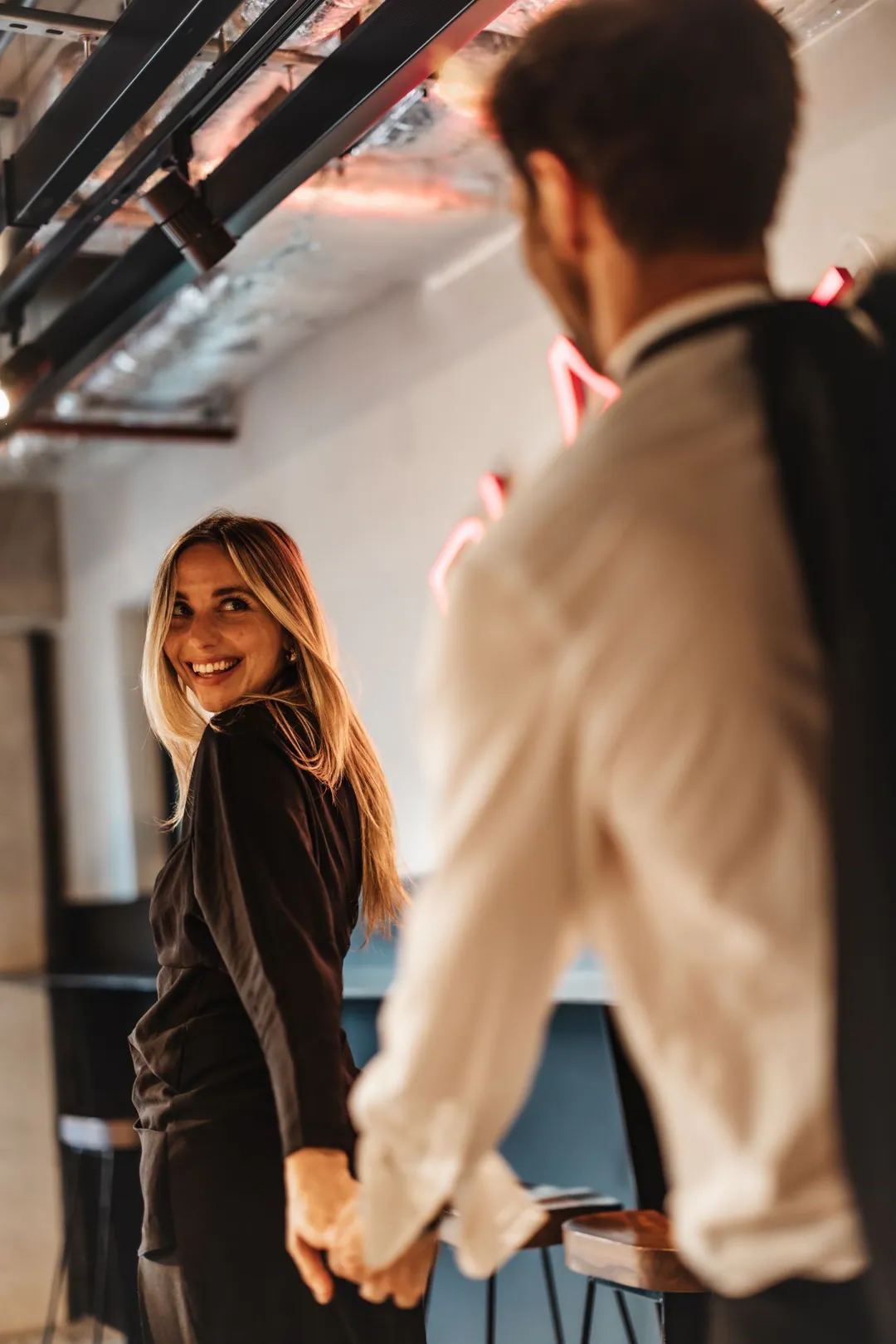 Woman in black smiling back at a man in a cream shirt as they hold hands indoors