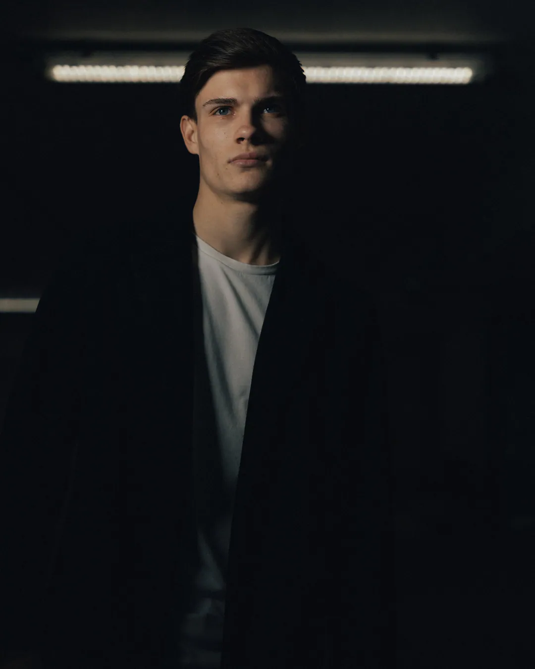 Man in a dark coat and white T-shirt standing in a dim car park, strip light glowing behind