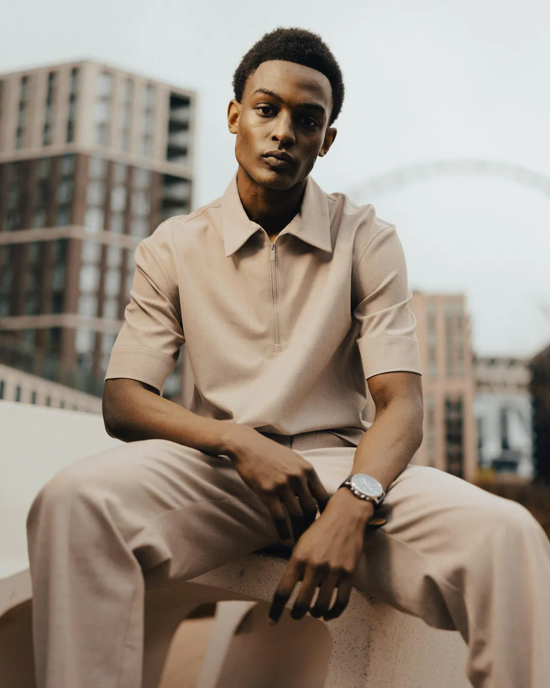 Man in a beige short-sleeved shirt and matching trousers seated on a stone bench, looking at the camera