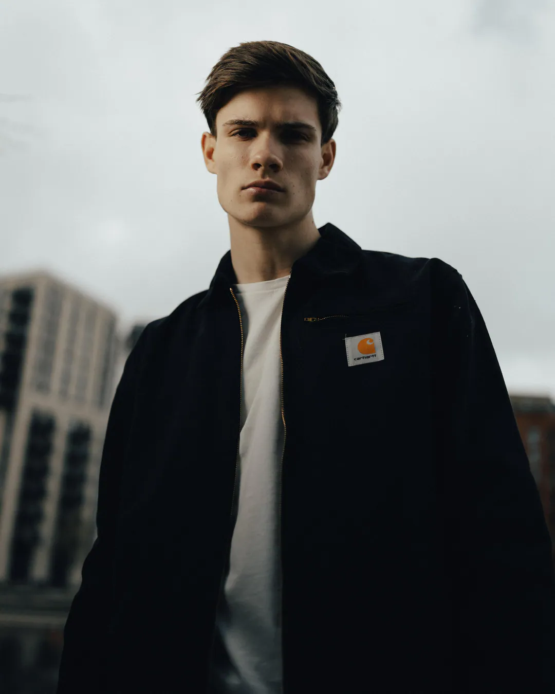 Man in a black workwear jacket and white T-shirt looking down at the camera under an overcast sky