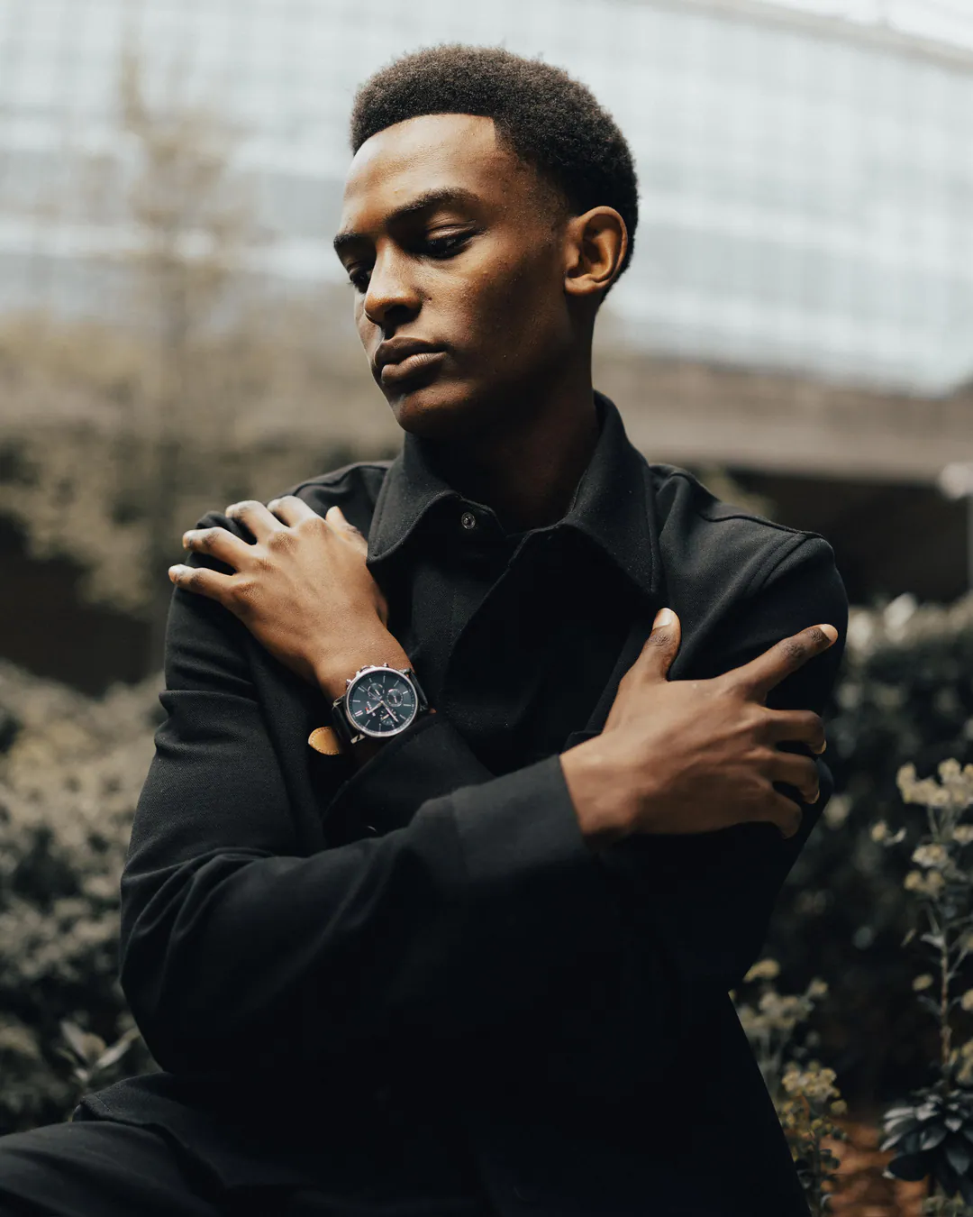 Man in a black shirt with arms crossed over his chest and a wristwatch, glass building behind