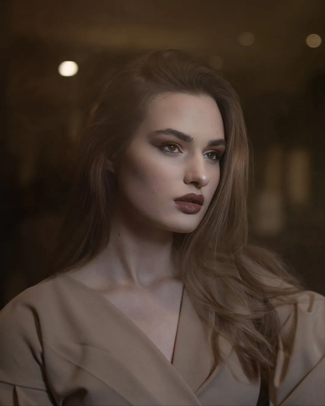 Woman with long brown hair in a beige wrap top gazing to one side in dim, warm light