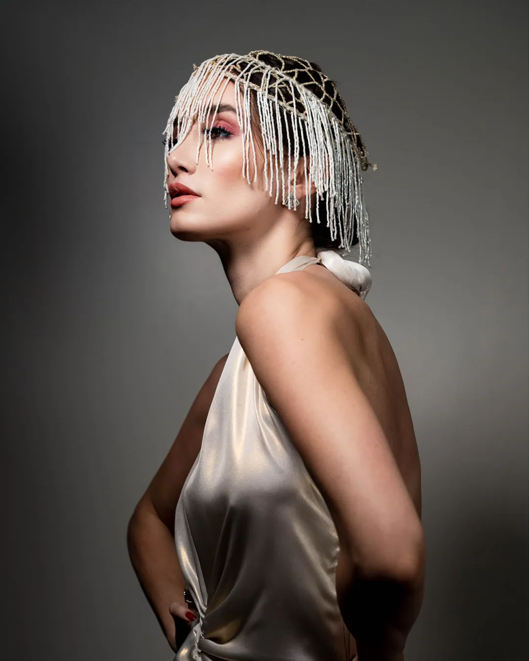Woman in profile wearing an ivory satin halter dress and beaded fringe headpiece against a grey backdrop