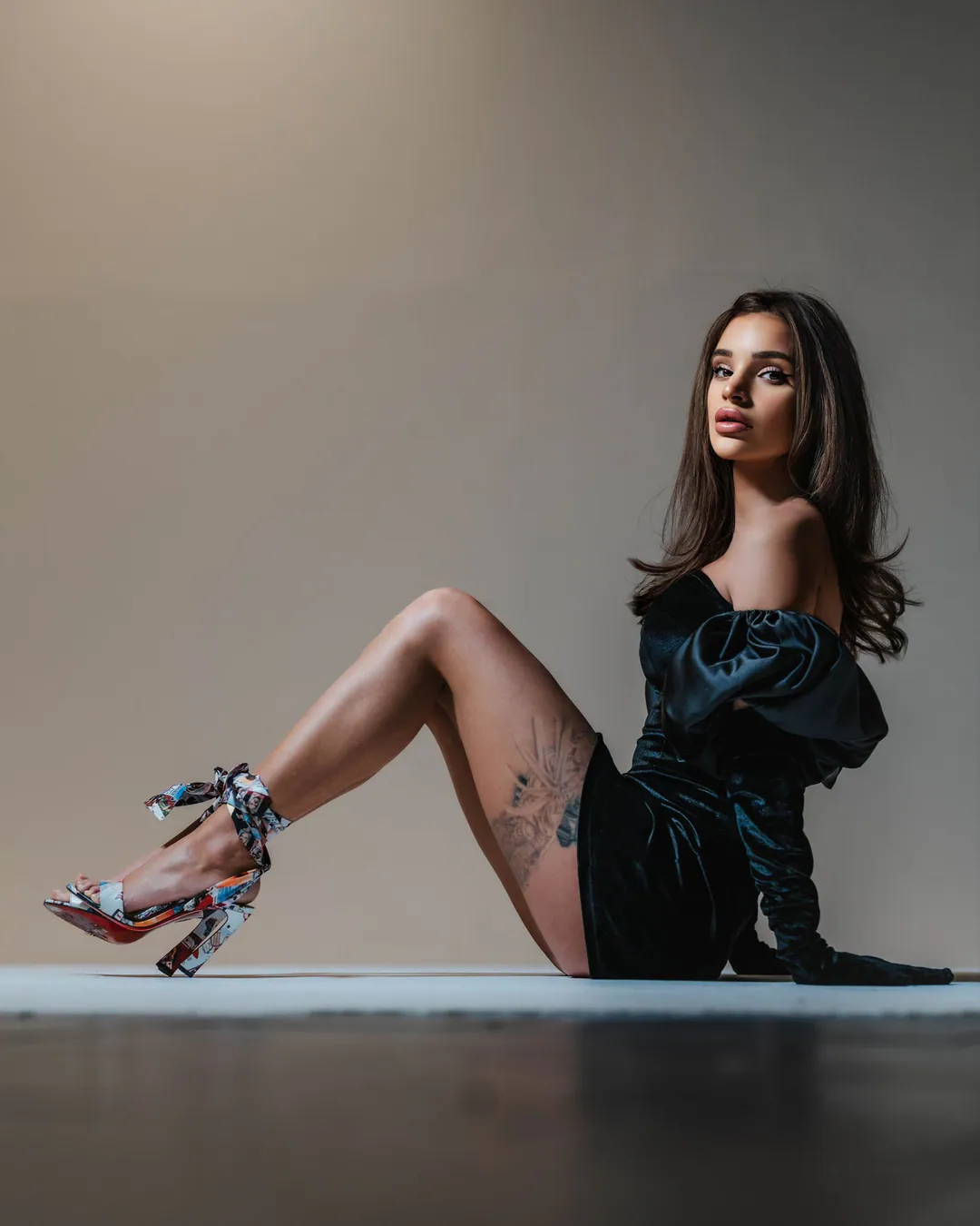 Woman in a black satin off-shoulder dress sitting on a studio floor with one knee raised, looking up