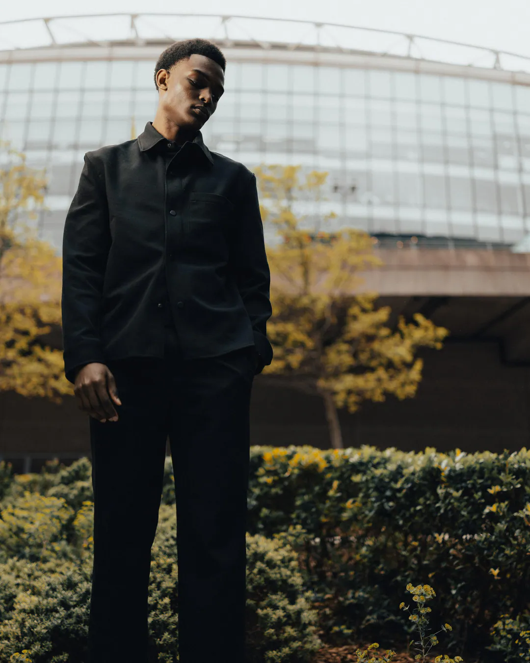 Man in a black overshirt and trousers standing among hedges and autumn trees before a curved glass building