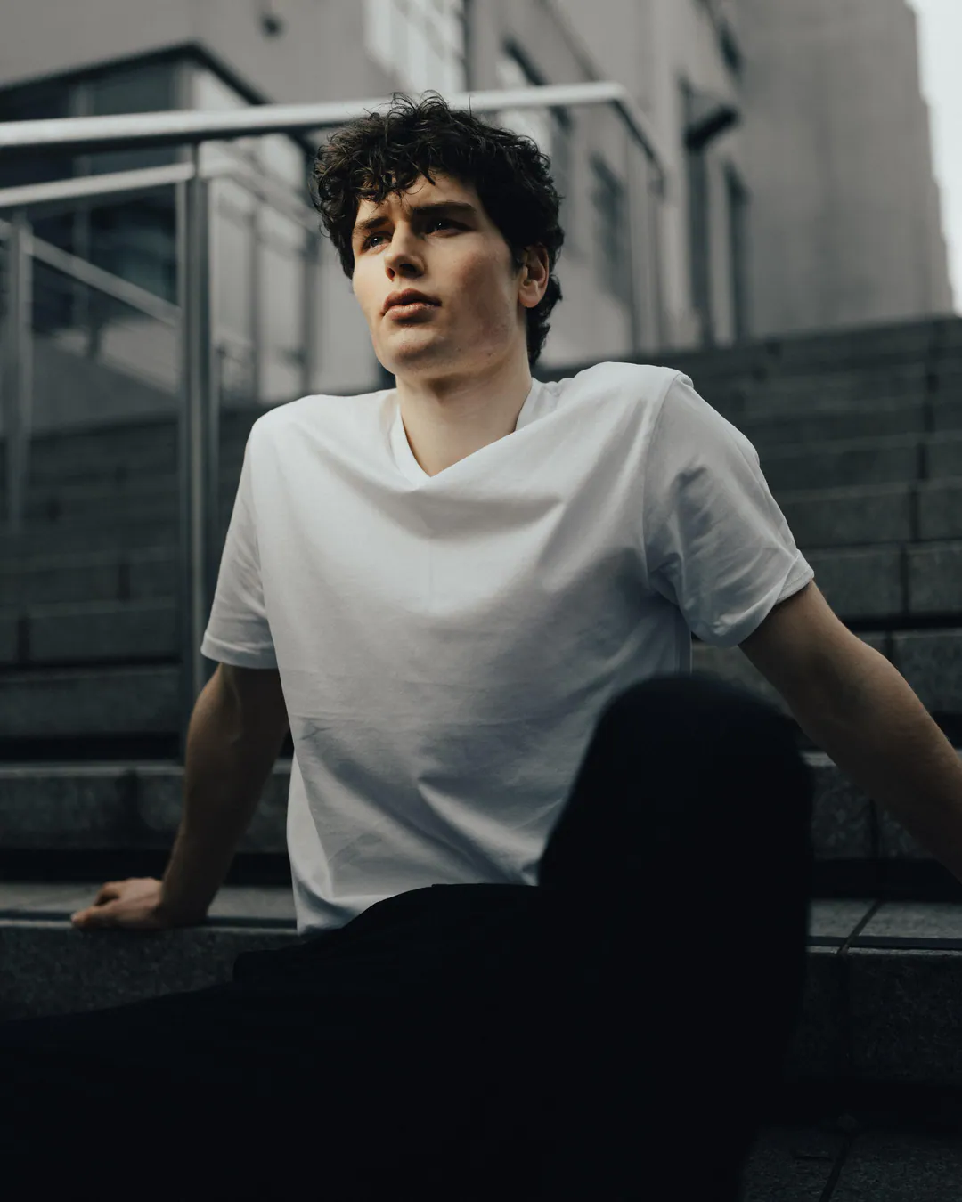 Man in a white V-neck T-shirt sitting on concrete steps beside a metal railing, gazing upwards