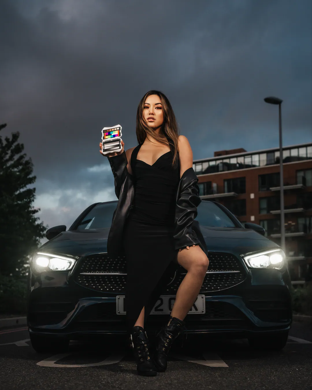 Woman in a black dress and leather jacket before a car with glowing headlights under stormy skies