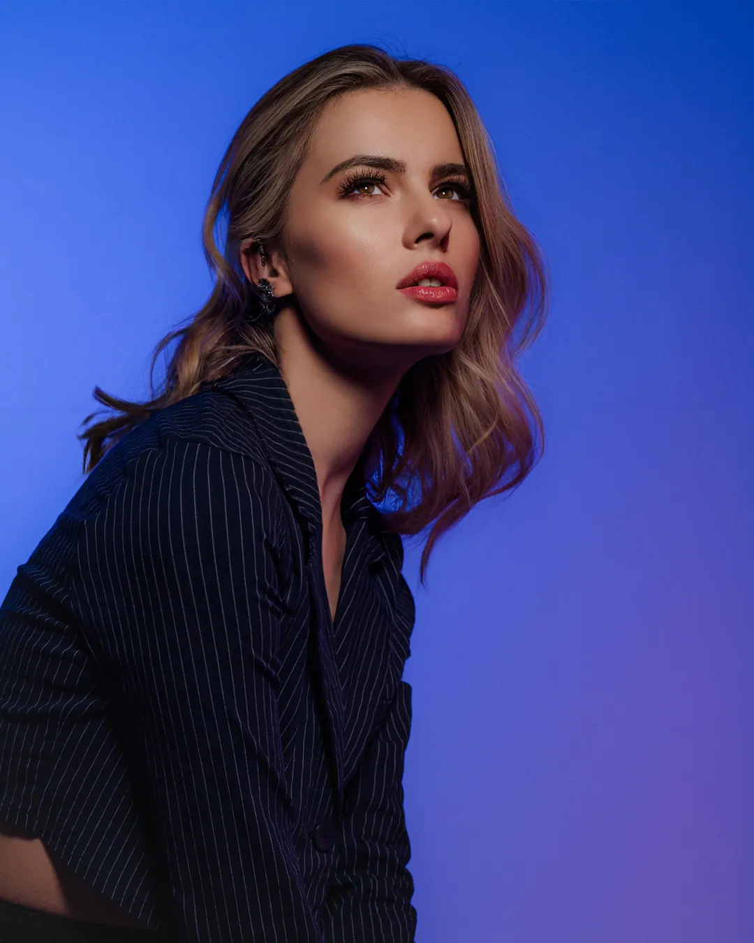 Woman with wavy hair in a pinstripe blazer looking upward against a vivid blue backdrop