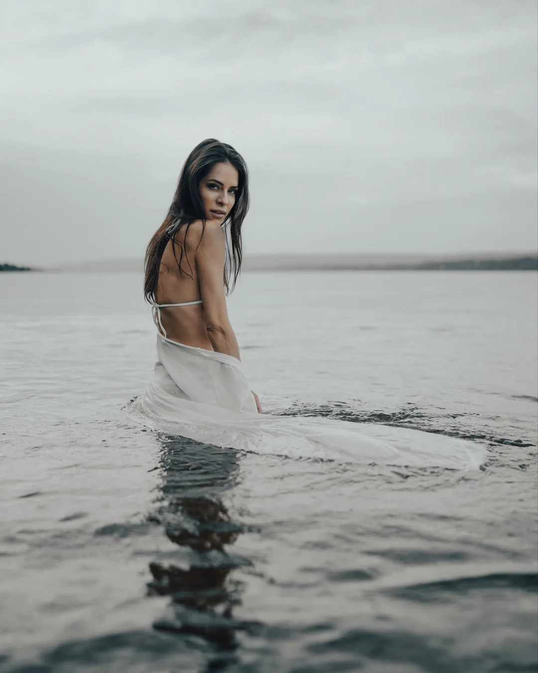 Woman in a sheer white wrap wading through calm grey water, looking back over her shoulder