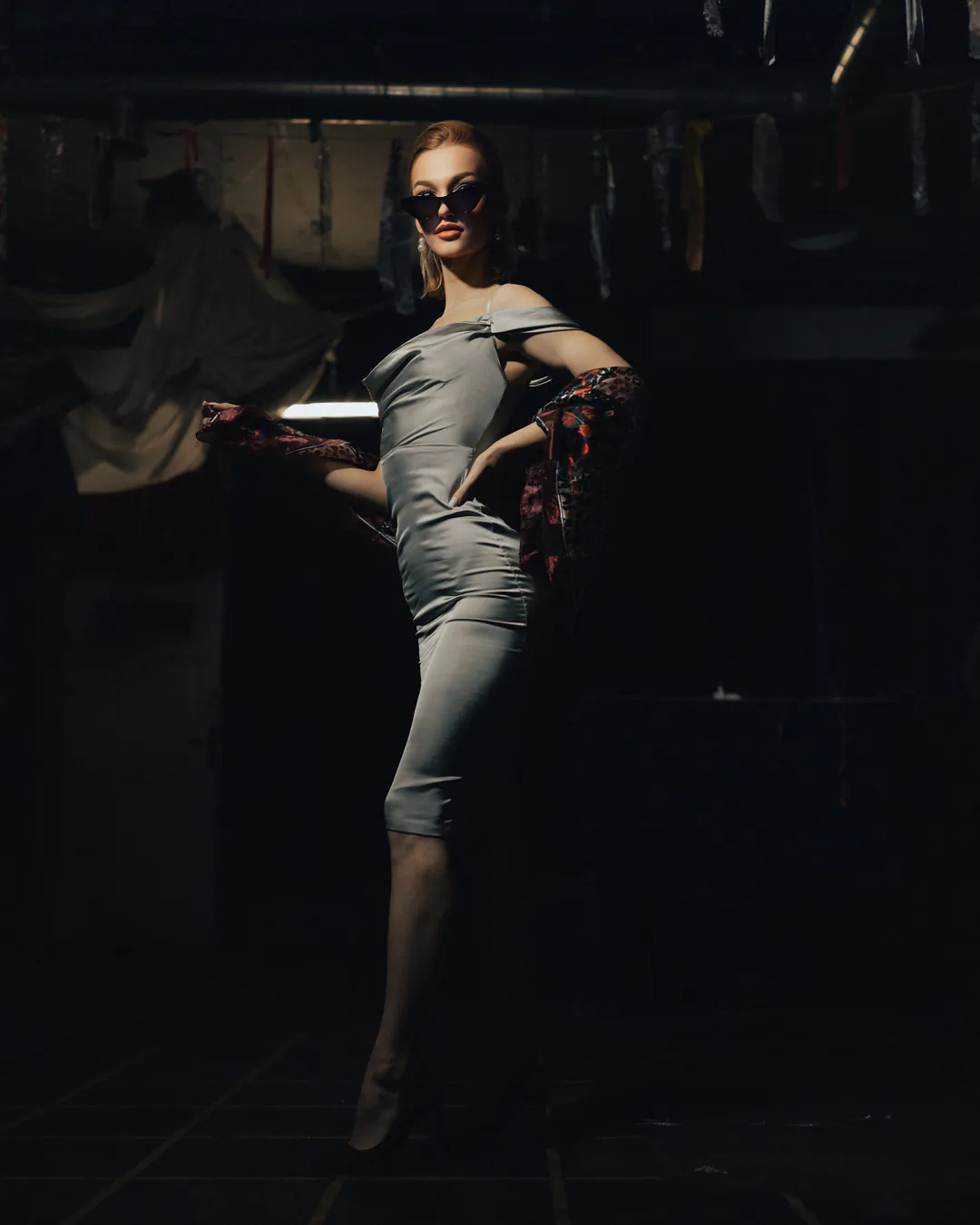 Woman in a fitted silver satin dress and sunglasses posing in a dim, cluttered industrial room