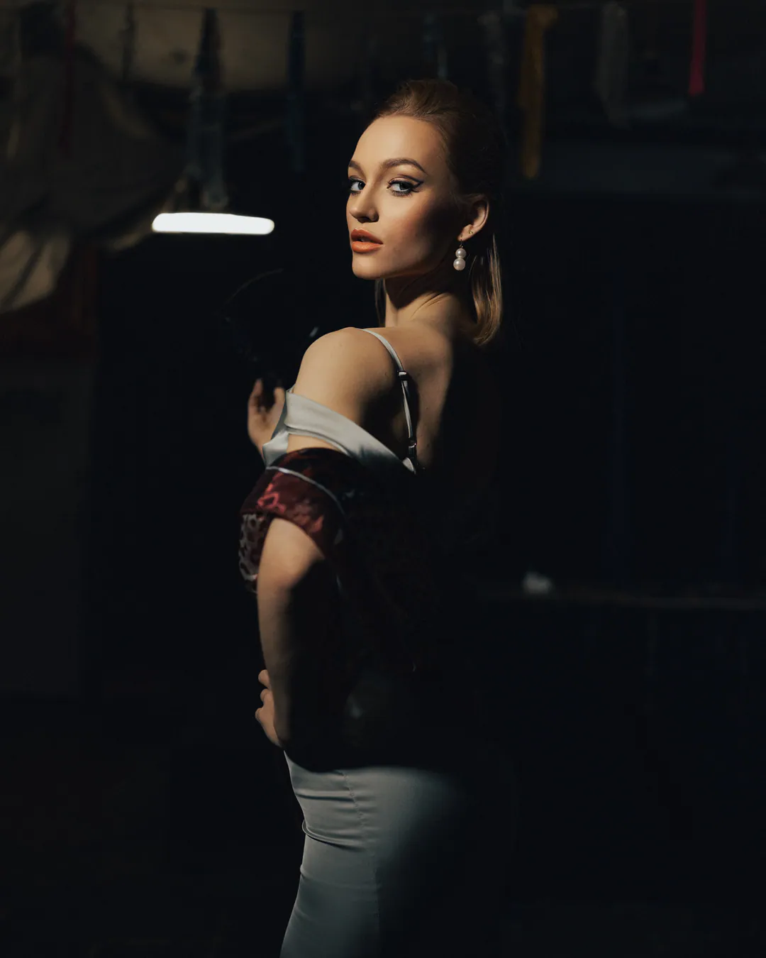 Woman in a satin dress glancing over her shoulder in a dark room lit by a strip light