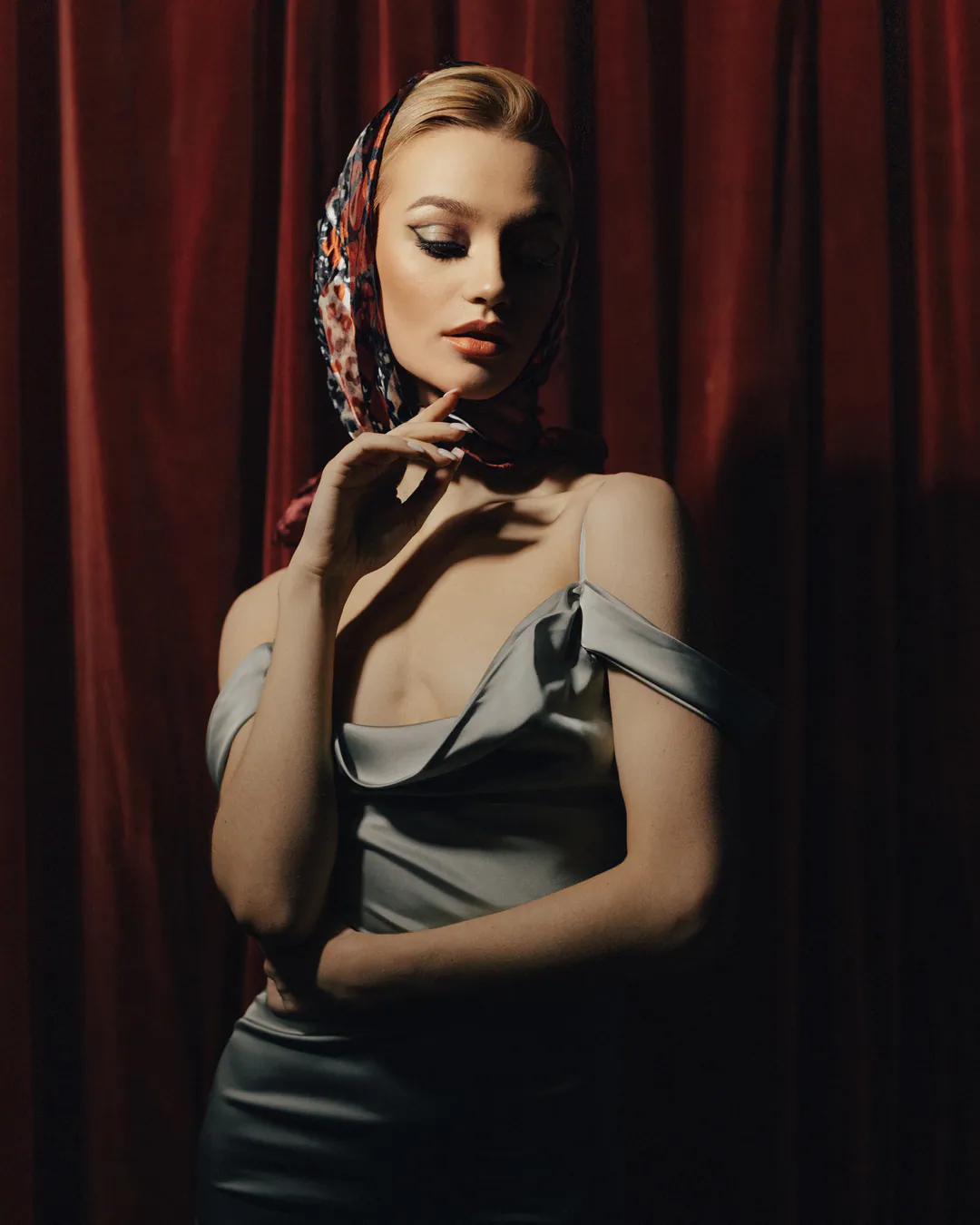 Woman in a silk headscarf and satin dress, hand by her chin, against a red velvet curtain