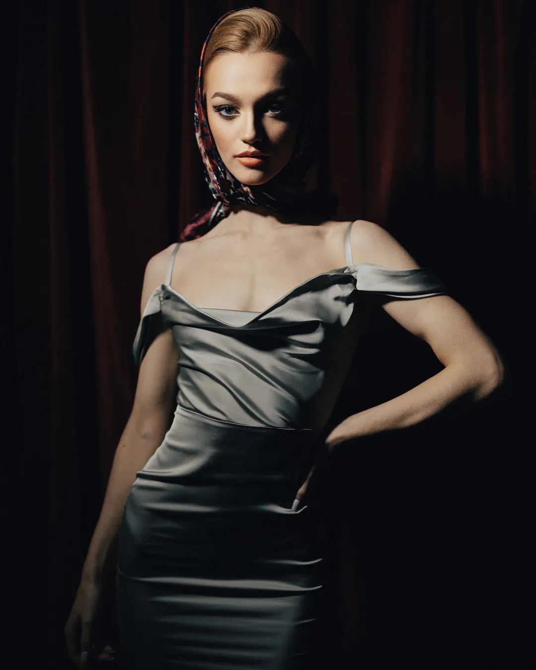 Woman in a silver satin dress and headscarf posing with one hand on her hip, side-lit against darkness
