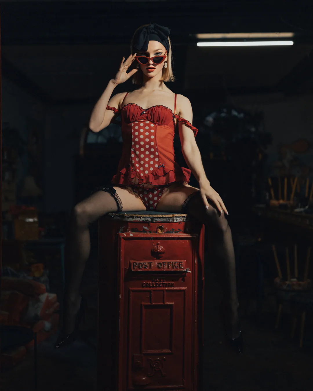 Woman in a red polka-dot corset and stockings lowering her sunglasses while seated on a red post box