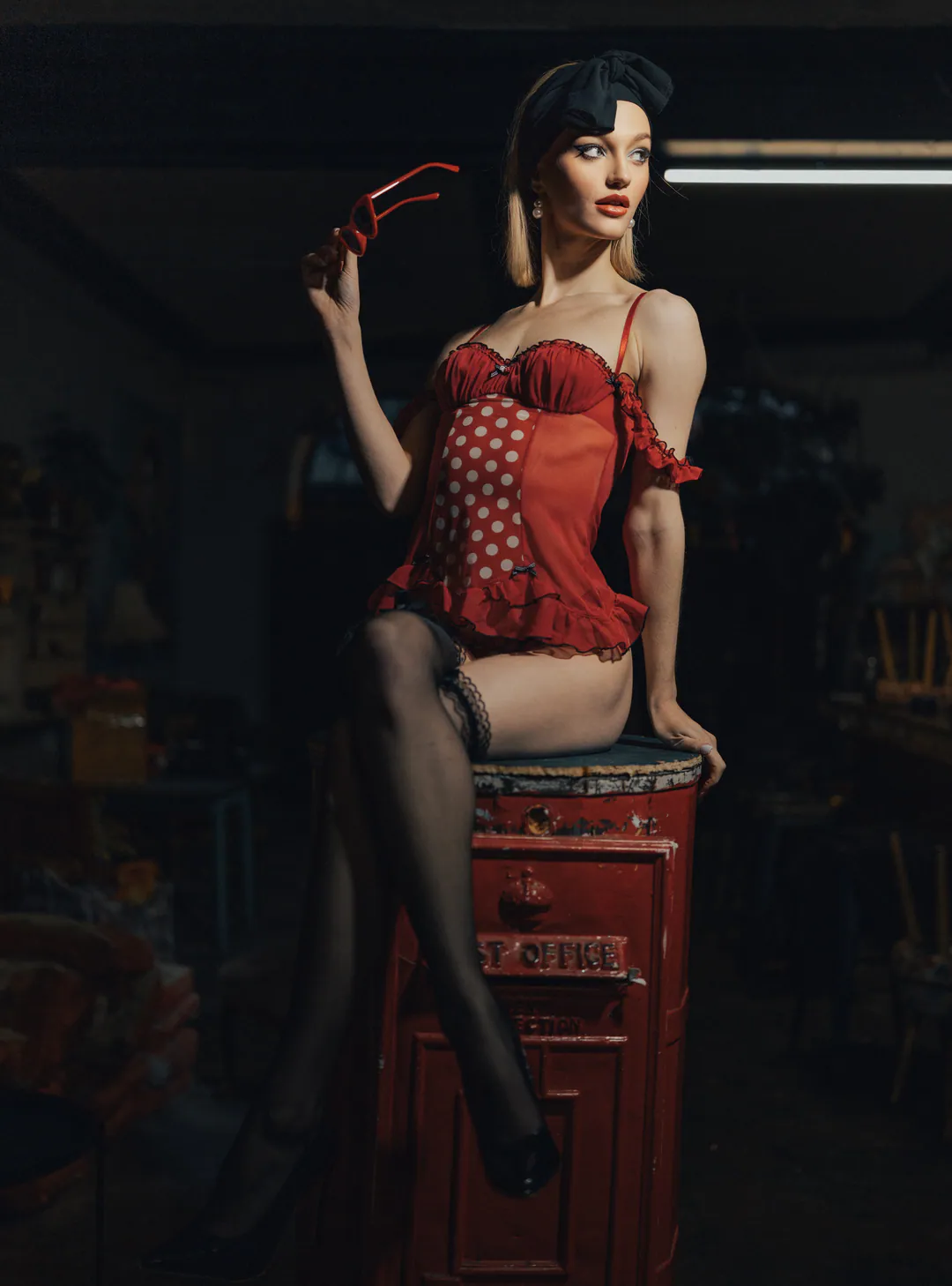 Woman in a red polka-dot corset and stockings perched on a red post box, holding sunglasses aloft