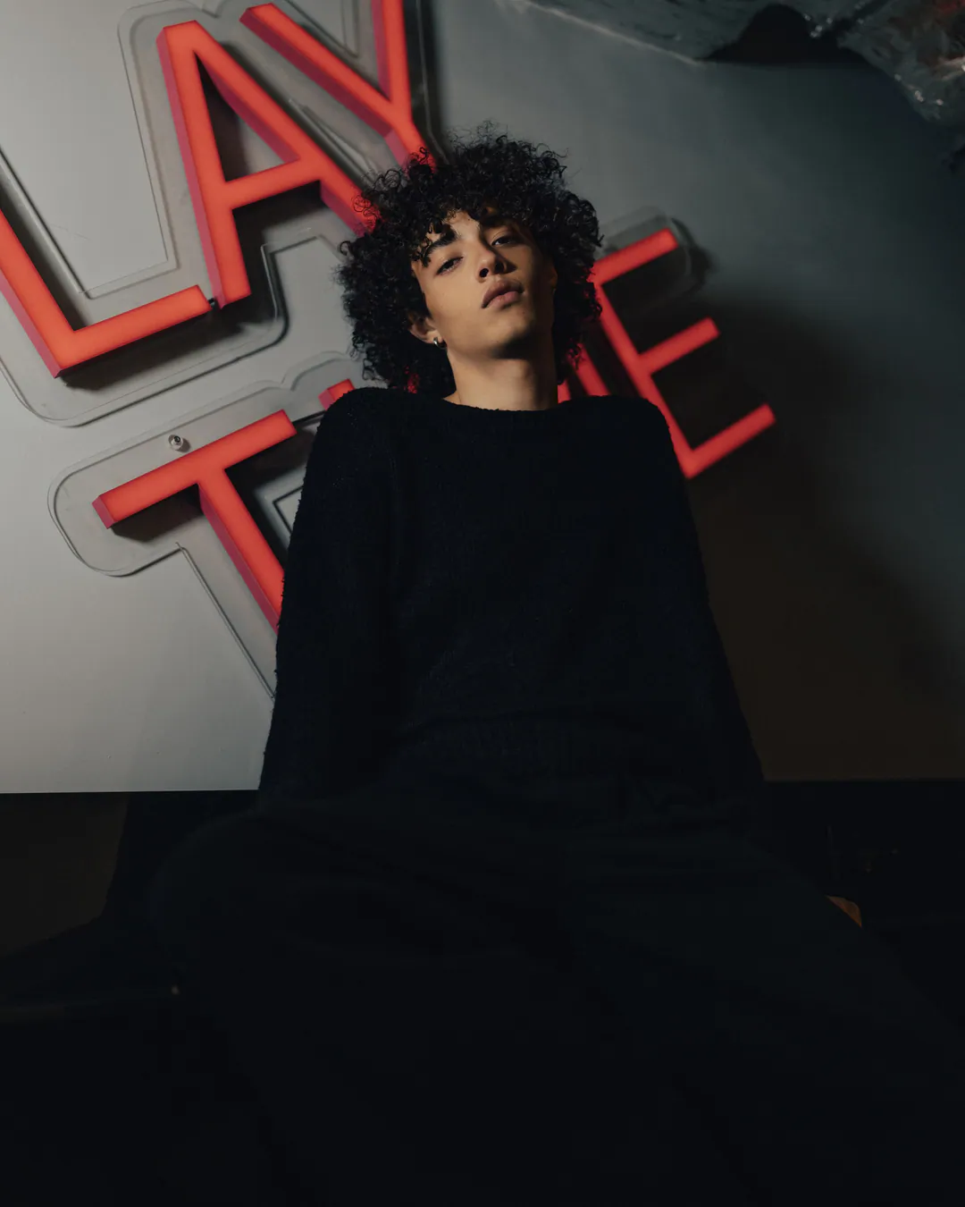 Person with curly hair in all black, eyes closed and head tilted beneath red neon lettering