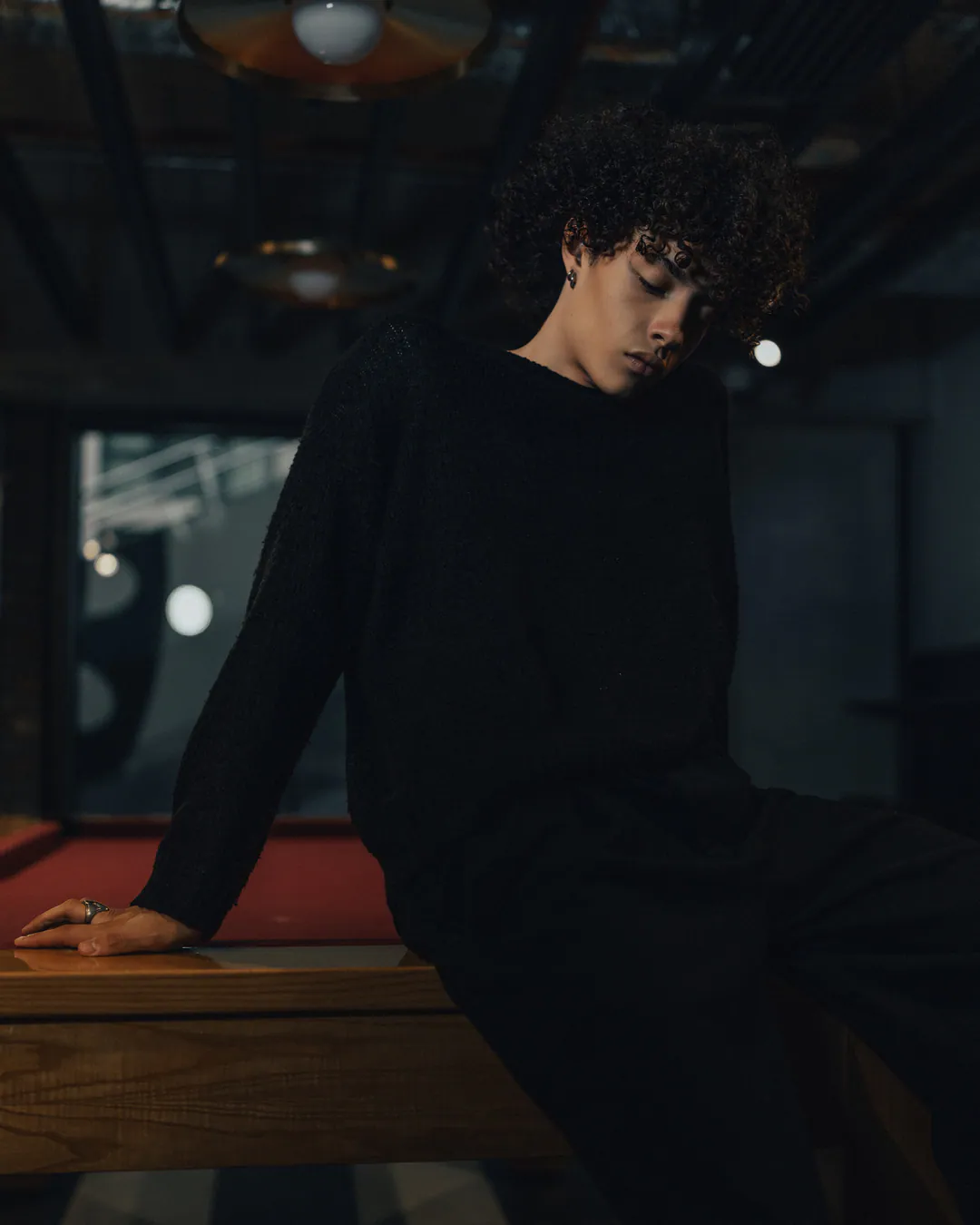 Person with curly hair in all black perched on the edge of a pool table, head bowed