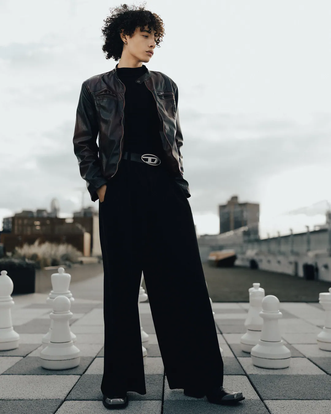 Person with curly hair in a burgundy leather jacket and wide black trousers among giant rooftop chess pieces
