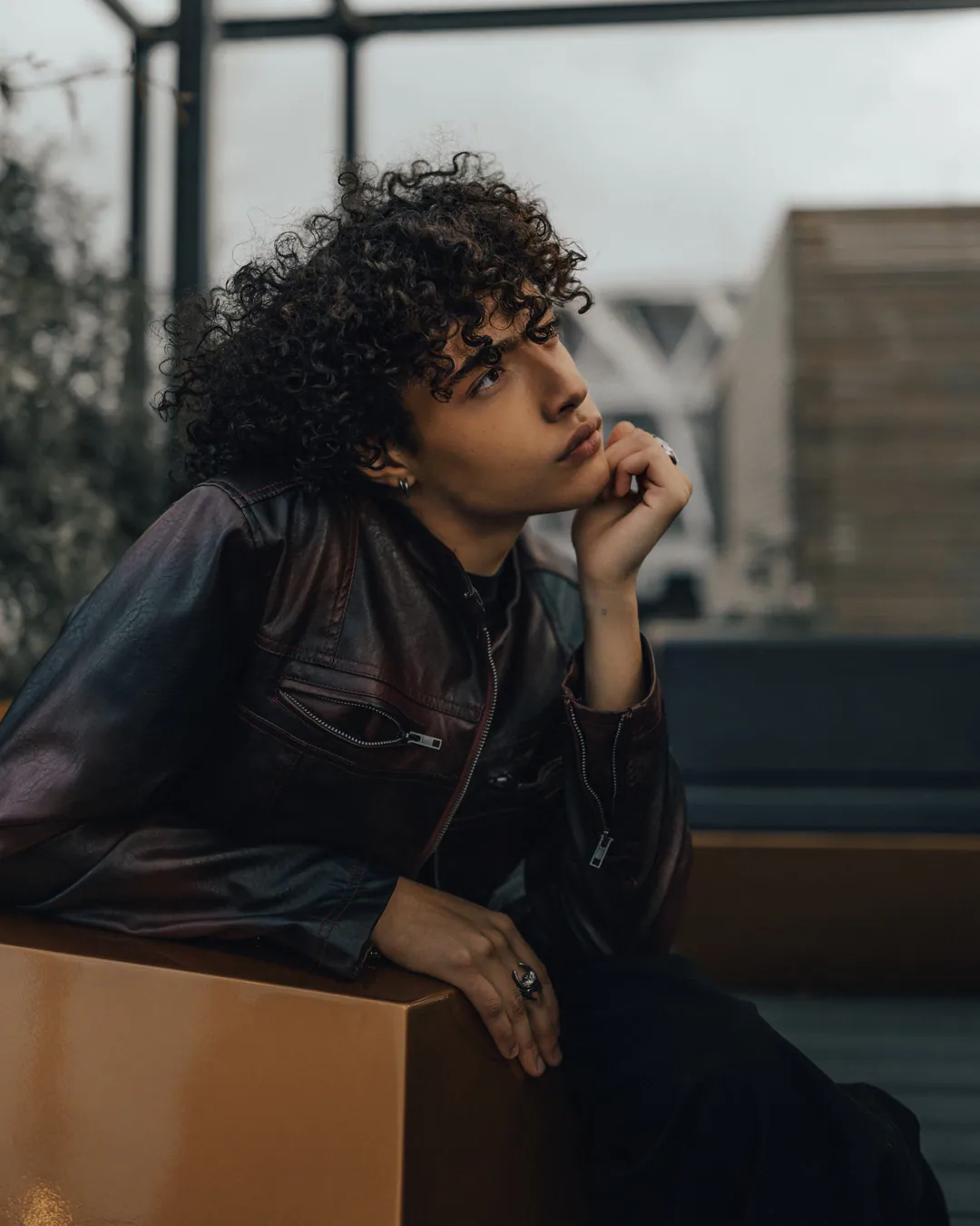 Person with curly hair in a leather jacket resting their chin on one hand against a rooftop sofa