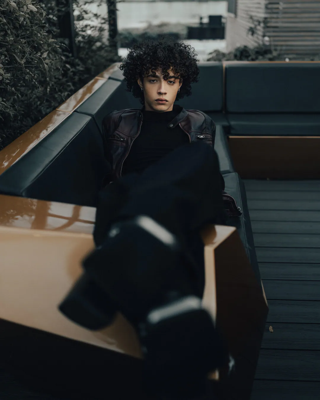 Person in a leather jacket reclining on a corner sofa with one boot raised towards the camera