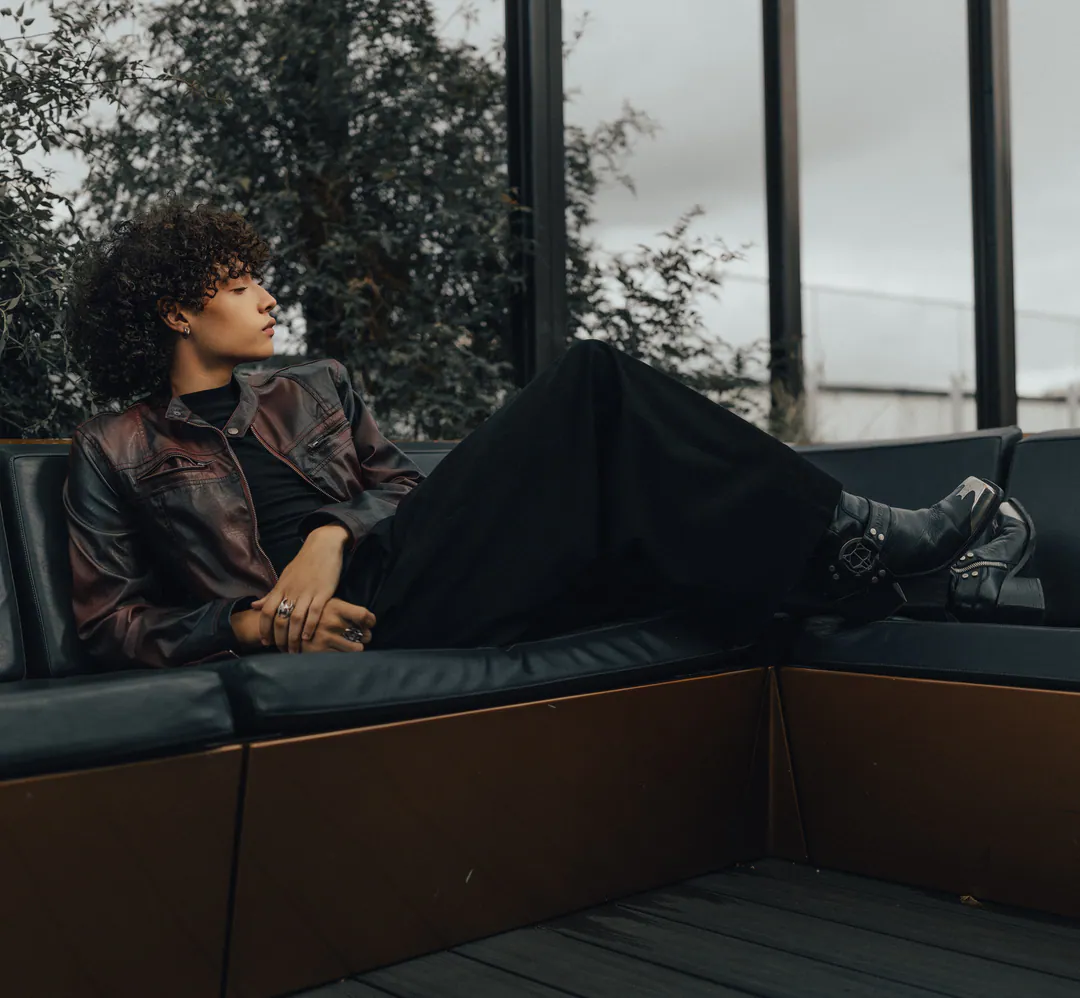 Person with curly hair in a leather jacket lounging full length along a dark rooftop sofa