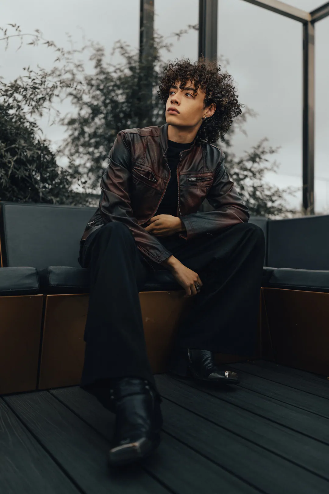 Person in a leather jacket and boots seated on a rooftop sofa, gazing upward beside greenery