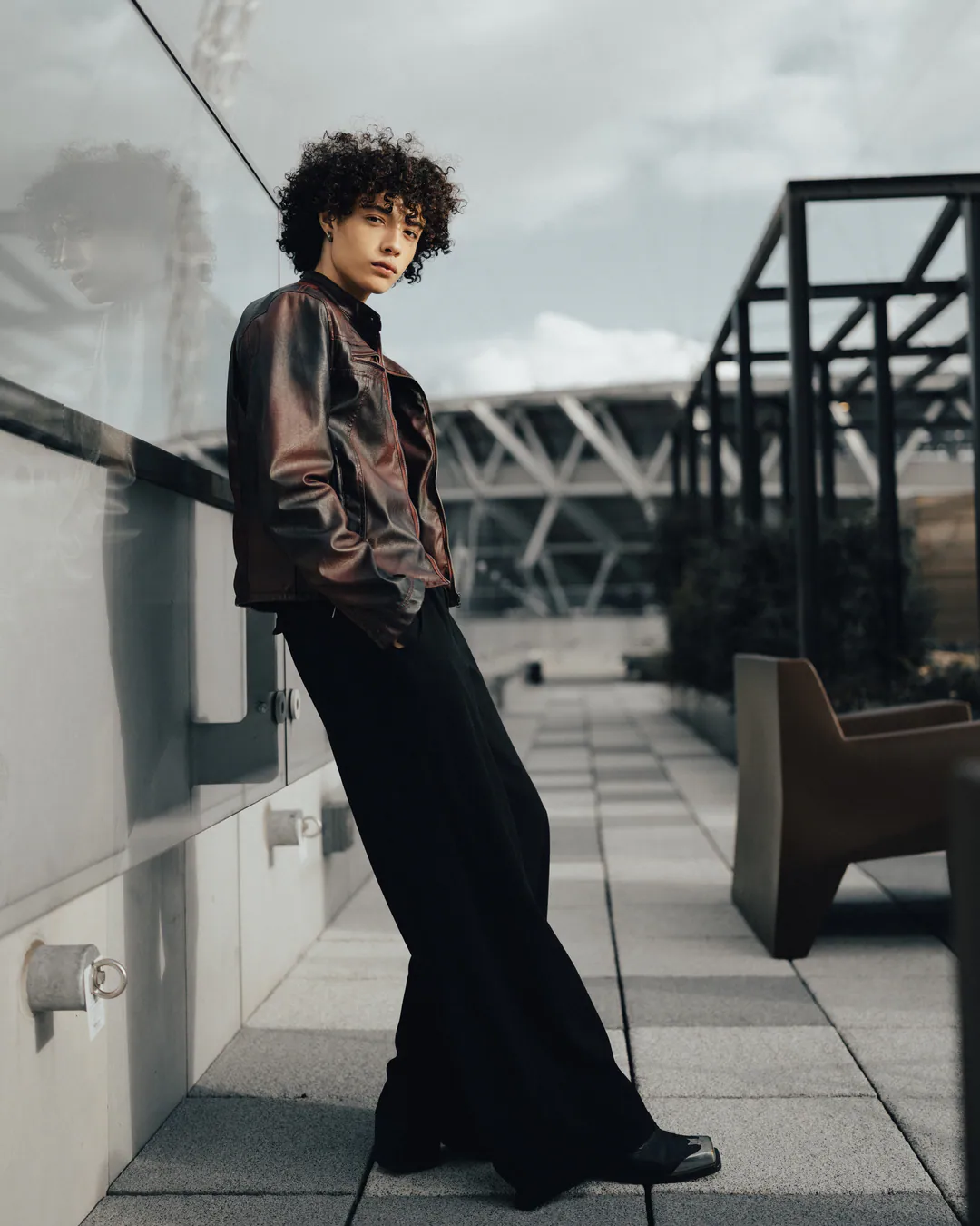 Person in a leather jacket and wide black trousers leaning against a glass balustrade on a rooftop terrace
