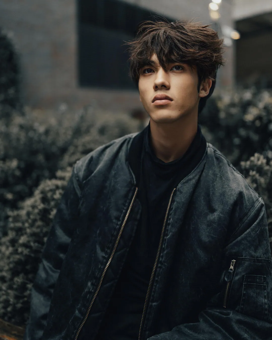 Man with tousled hair in a dark bomber jacket gazing upward beside hedges outdoors in muted light