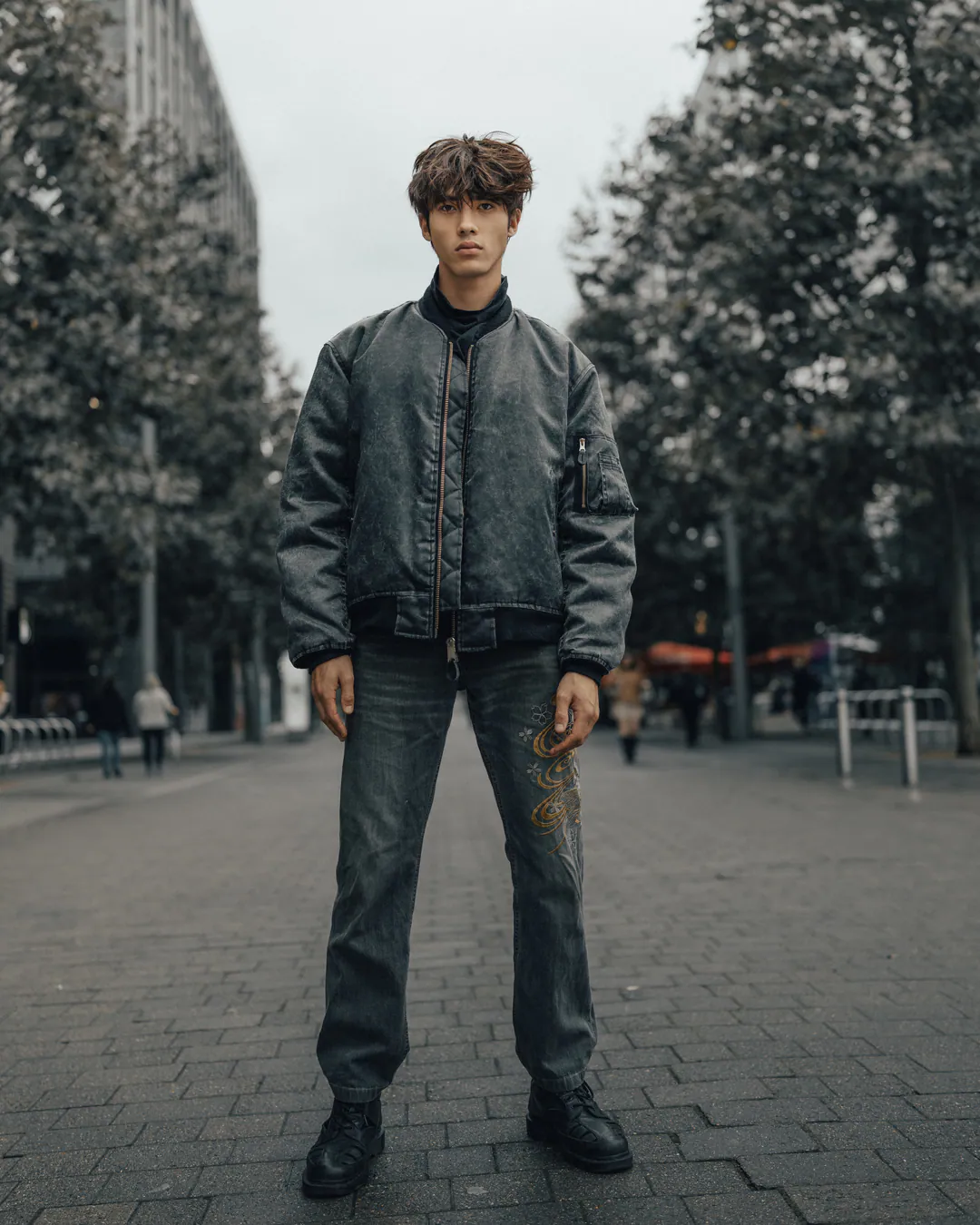 Man in a grey bomber jacket and jeans standing on a tree-lined paved street under overcast skies