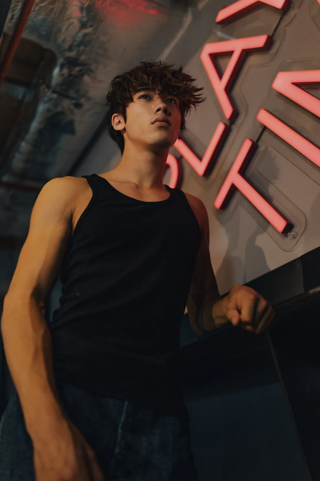 Man in a black vest top leaning on a bar beneath pink neon lettering, shot from below