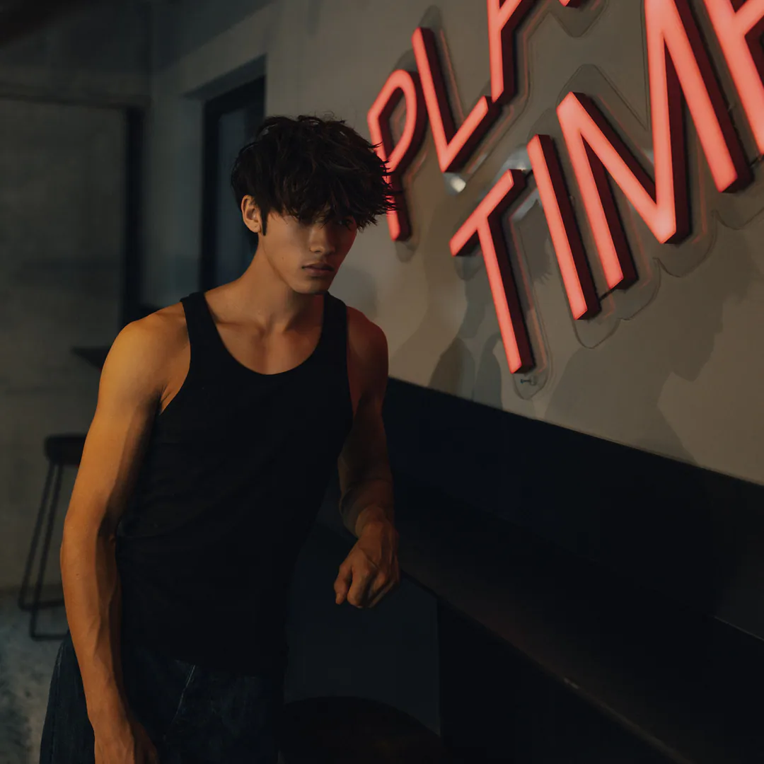 Man in a black vest top leaning against a dark bar counter beside large pink neon lettering