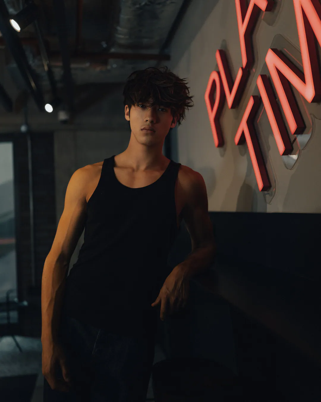 Man in a black vest top standing beside pink neon lettering in a dim bar with exposed ceiling
