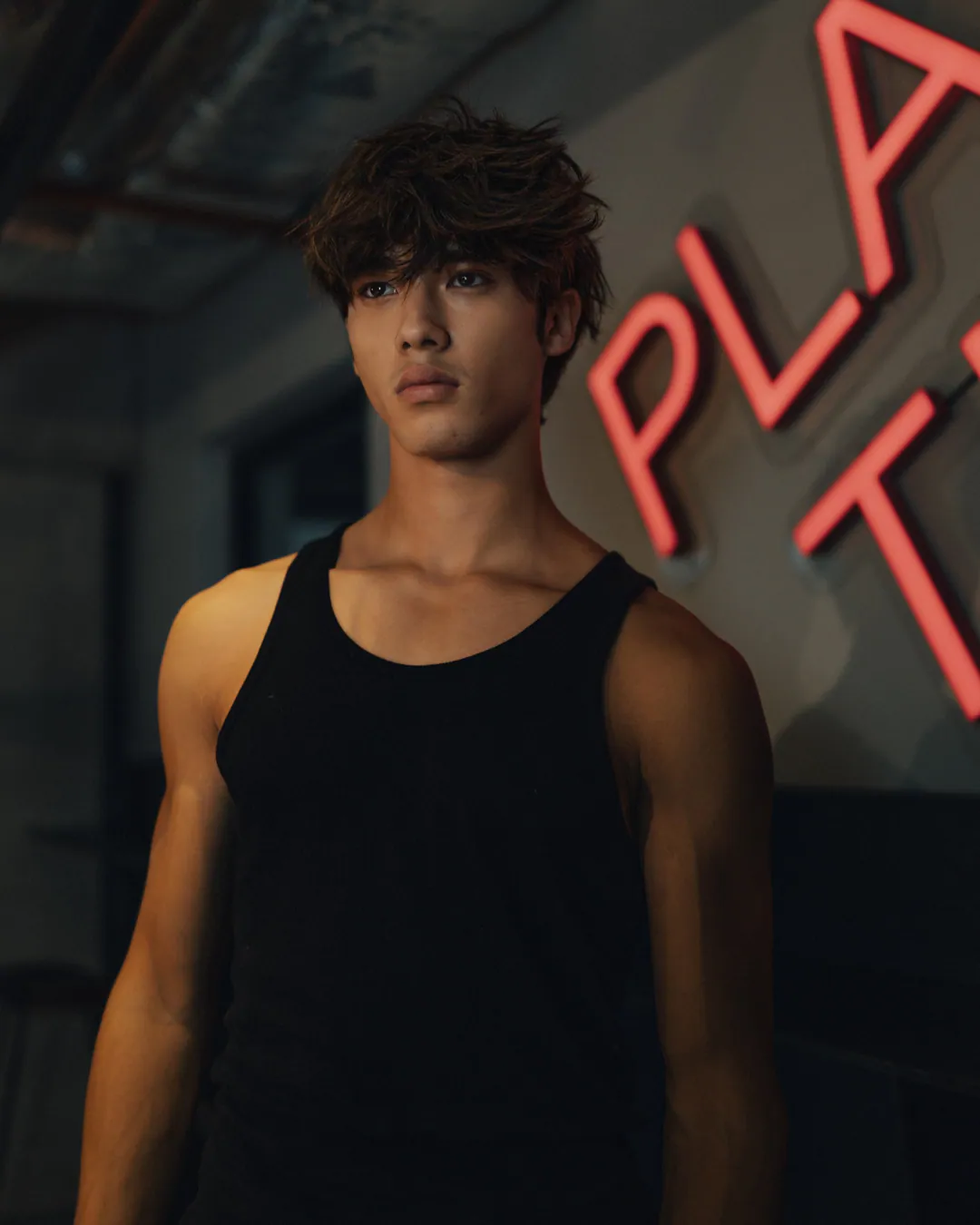 Man in a black vest top gazing upward in warm side light beside a pink neon sign