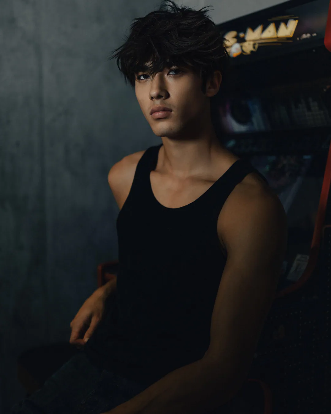 Man in a black vest top seated beside an arcade machine against a concrete wall in low light