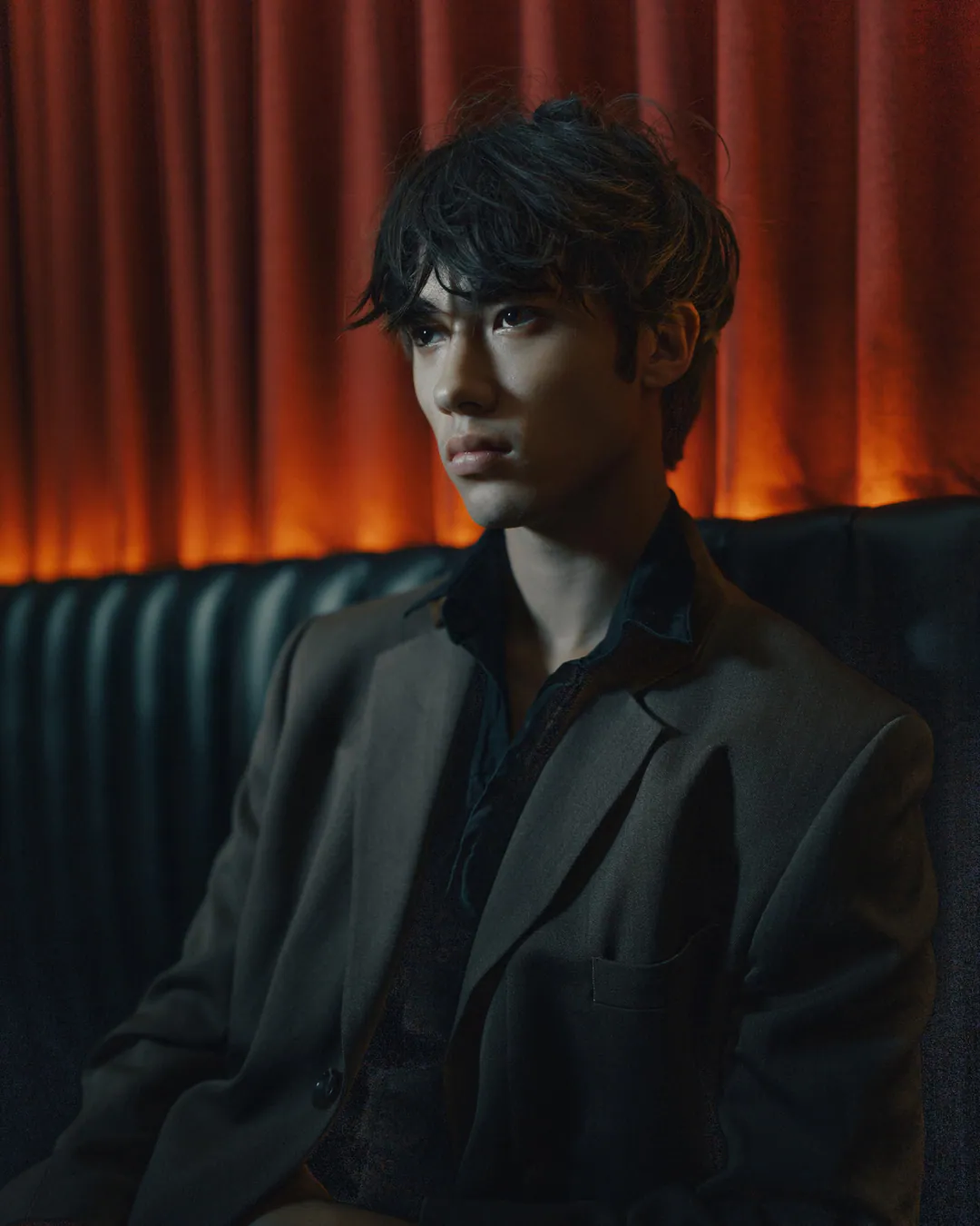 Man with tousled hair in a grey blazer gazing sideways in a leather booth beneath an orange-lit curtain