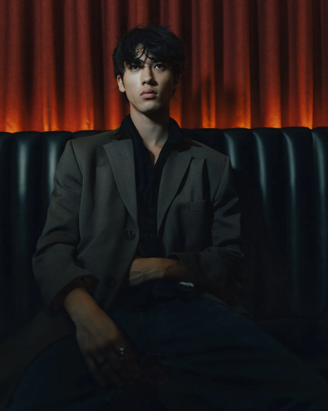 Man in a grey blazer lounging in a dark leather booth beneath a warm orange-lit curtain