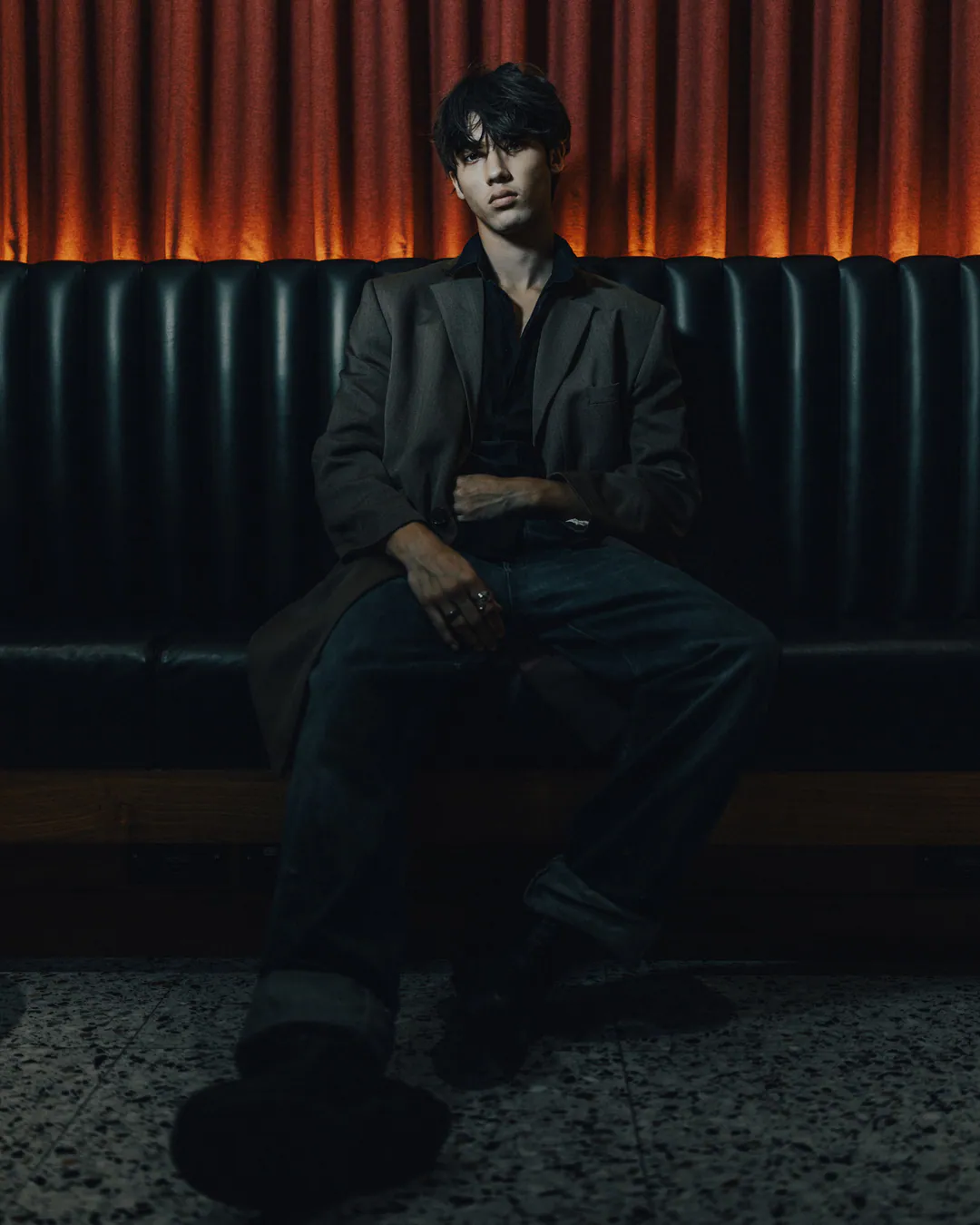 Man in a dark blazer and jeans seated with crossed legs on a leather banquette beneath orange light