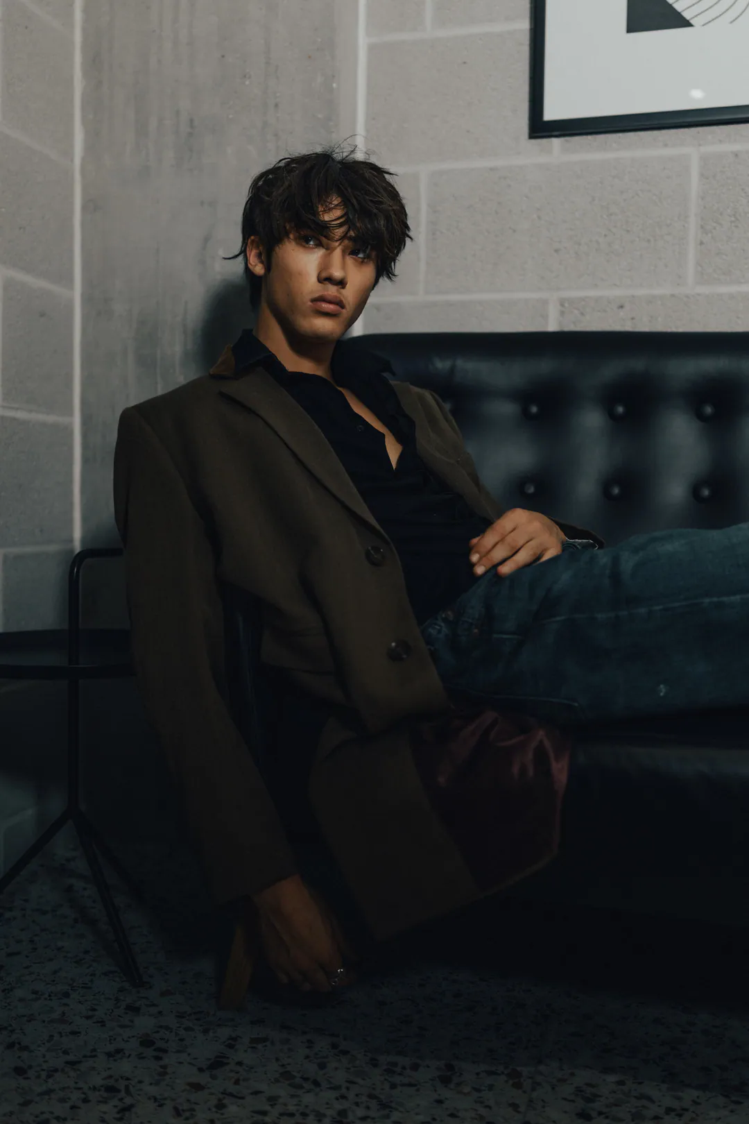 Man in an olive blazer reclining on a black leather sofa against a pale breeze-block wall