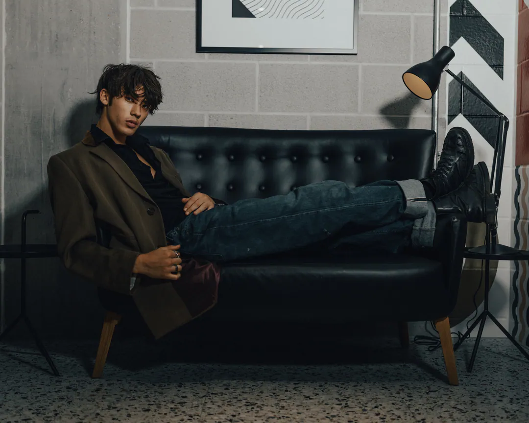 Man in an olive blazer and jeans stretched across a black tufted leather sofa beside a floor lamp