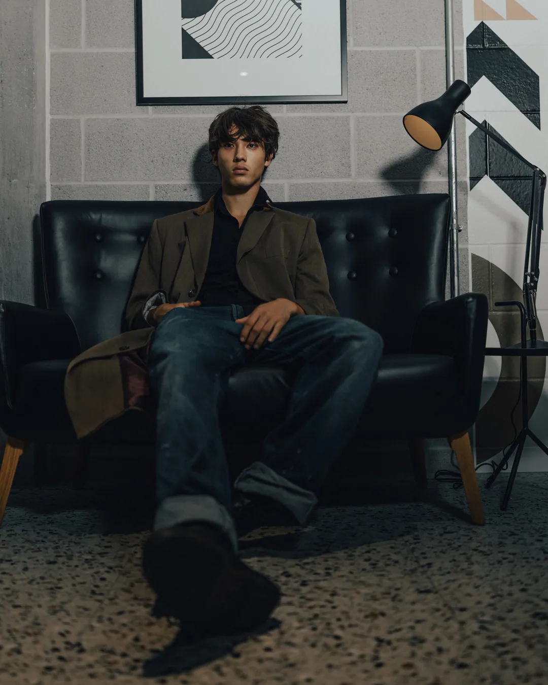 Man in an olive blazer sitting on a black leather sofa with one boot towards the camera