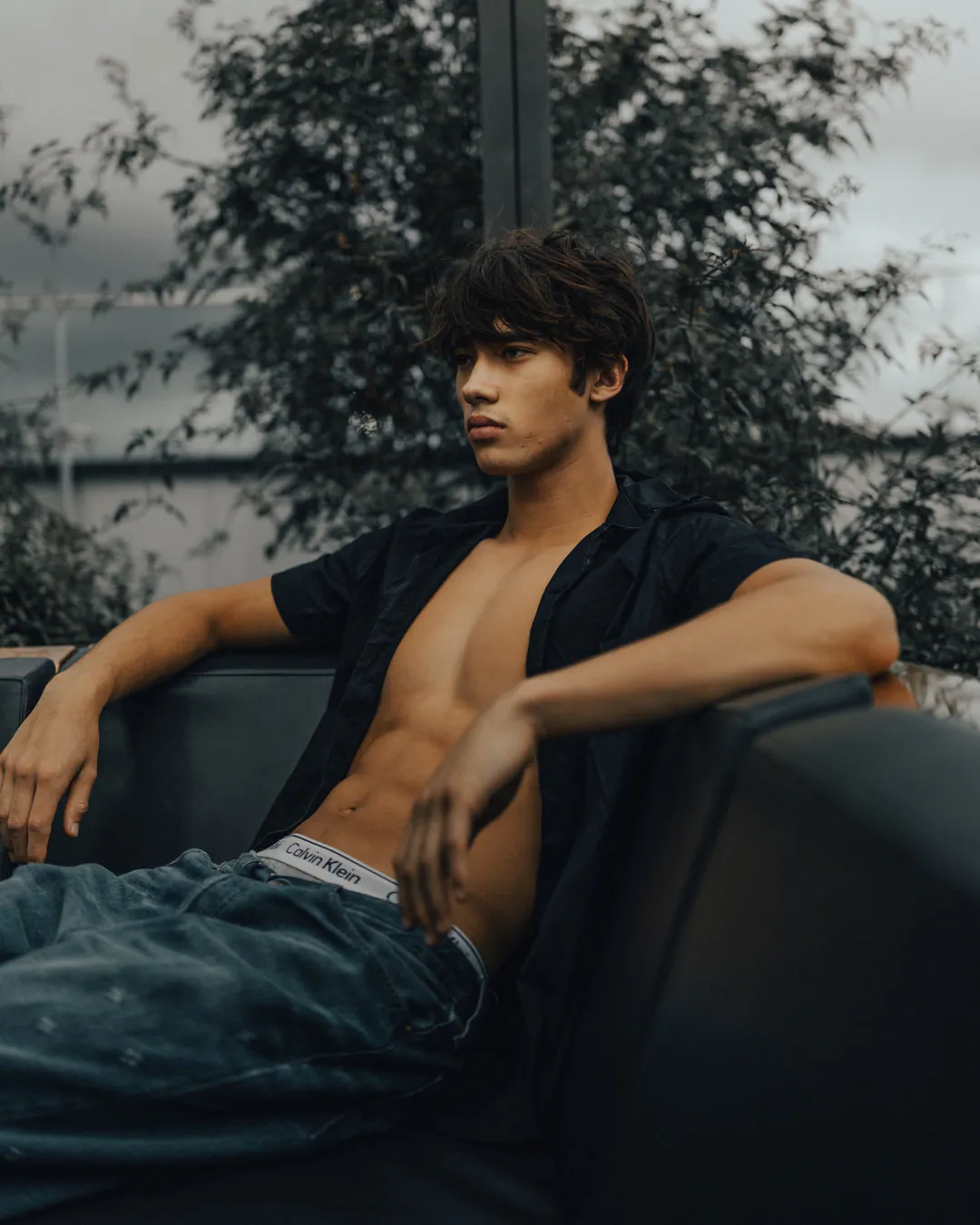 Man in an unbuttoned black shirt and jeans reclining on a dark sofa beside a leafy window