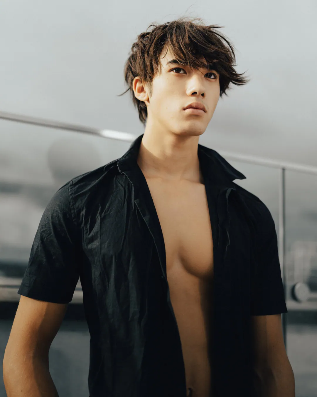 Man with tousled hair in an open black short-sleeved shirt on a rooftop under an overcast sky