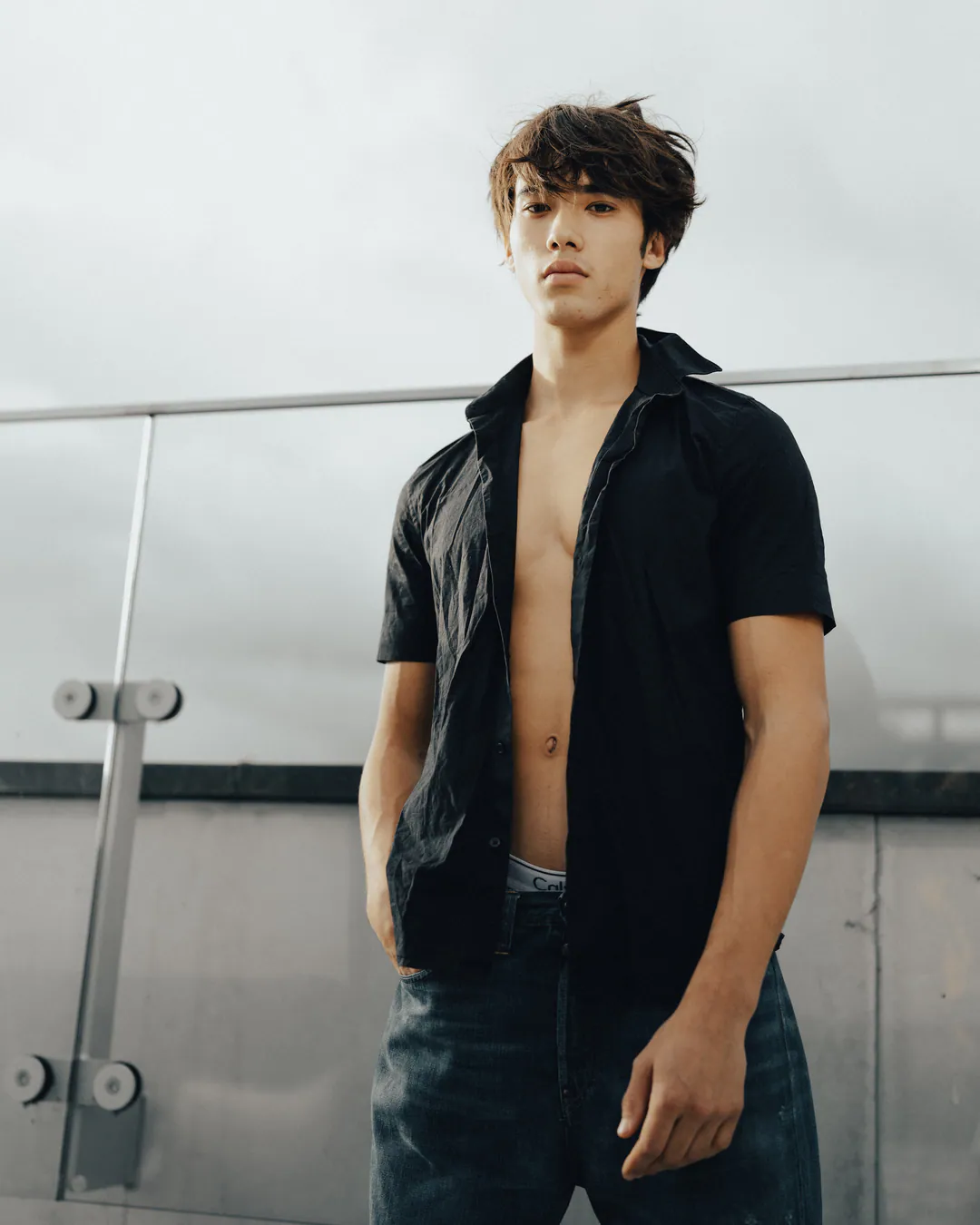 Man in an open black shirt and jeans standing by a glass balustrade on a grey rooftop