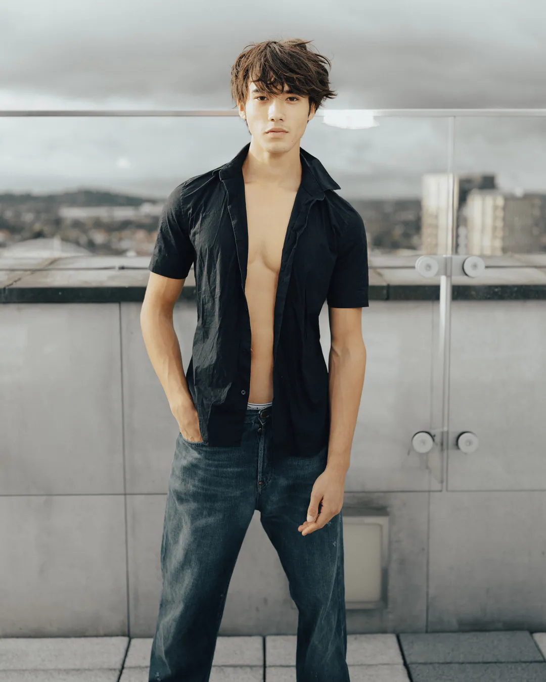Man in an open black shirt and jeans with hands in pockets on a rooftop overlooking city buildings