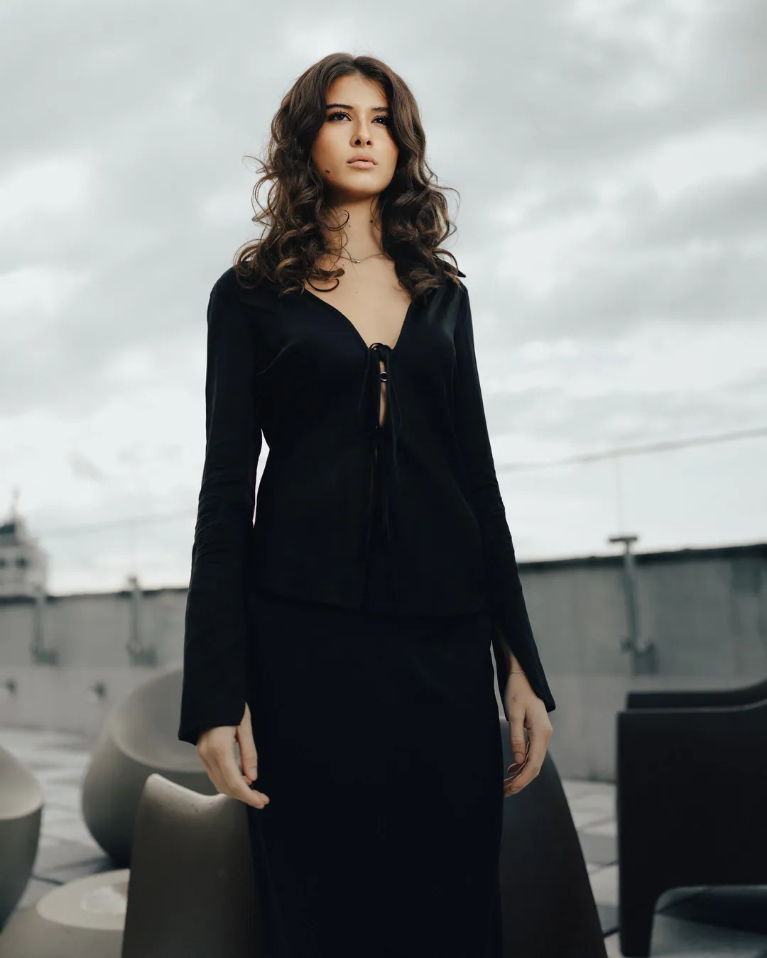 Woman in a black tie-front top and long skirt on a rooftop terrace under grey skies