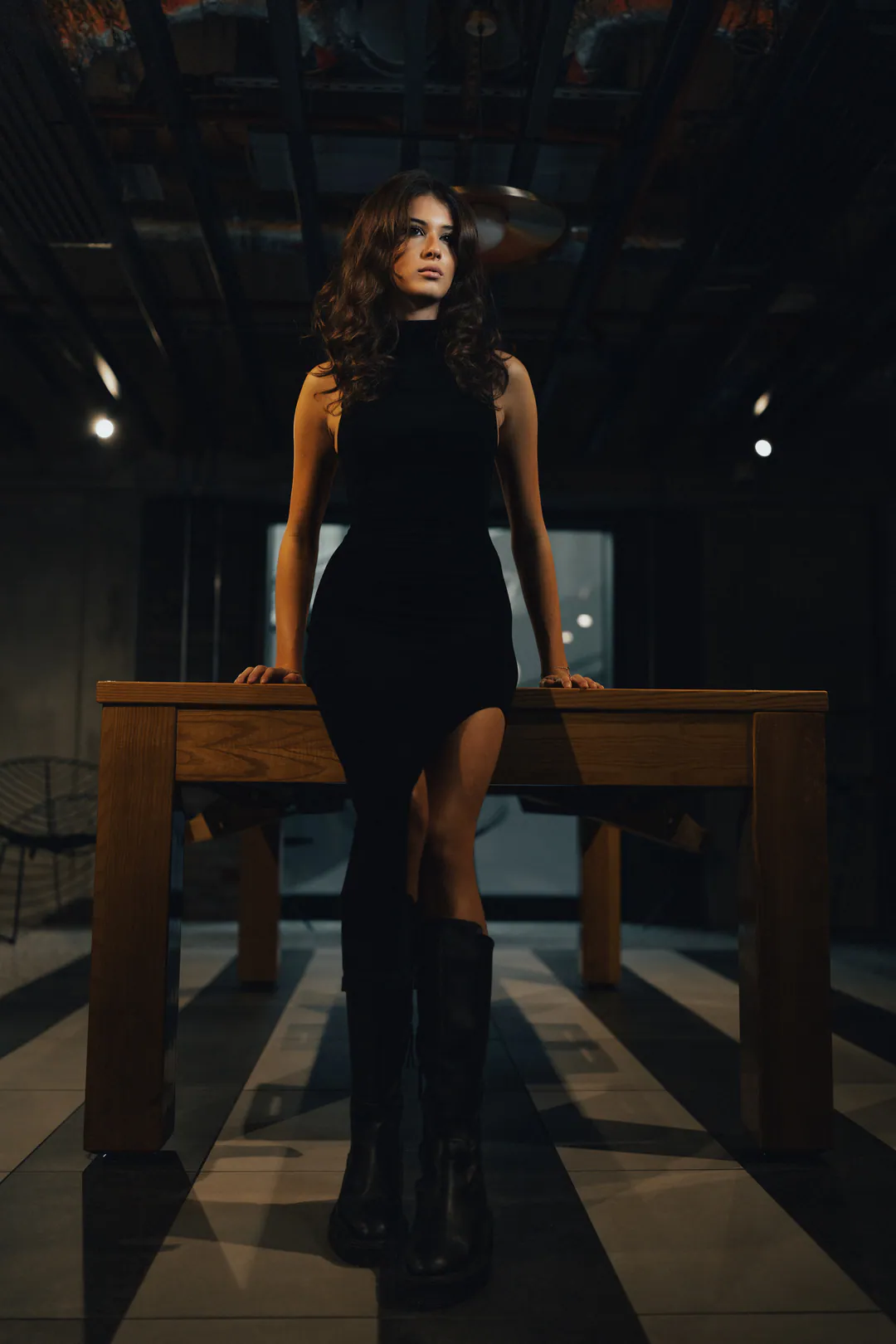 Woman in a black sleeveless dress and knee-high boots leaning against a wooden table in a dim bar