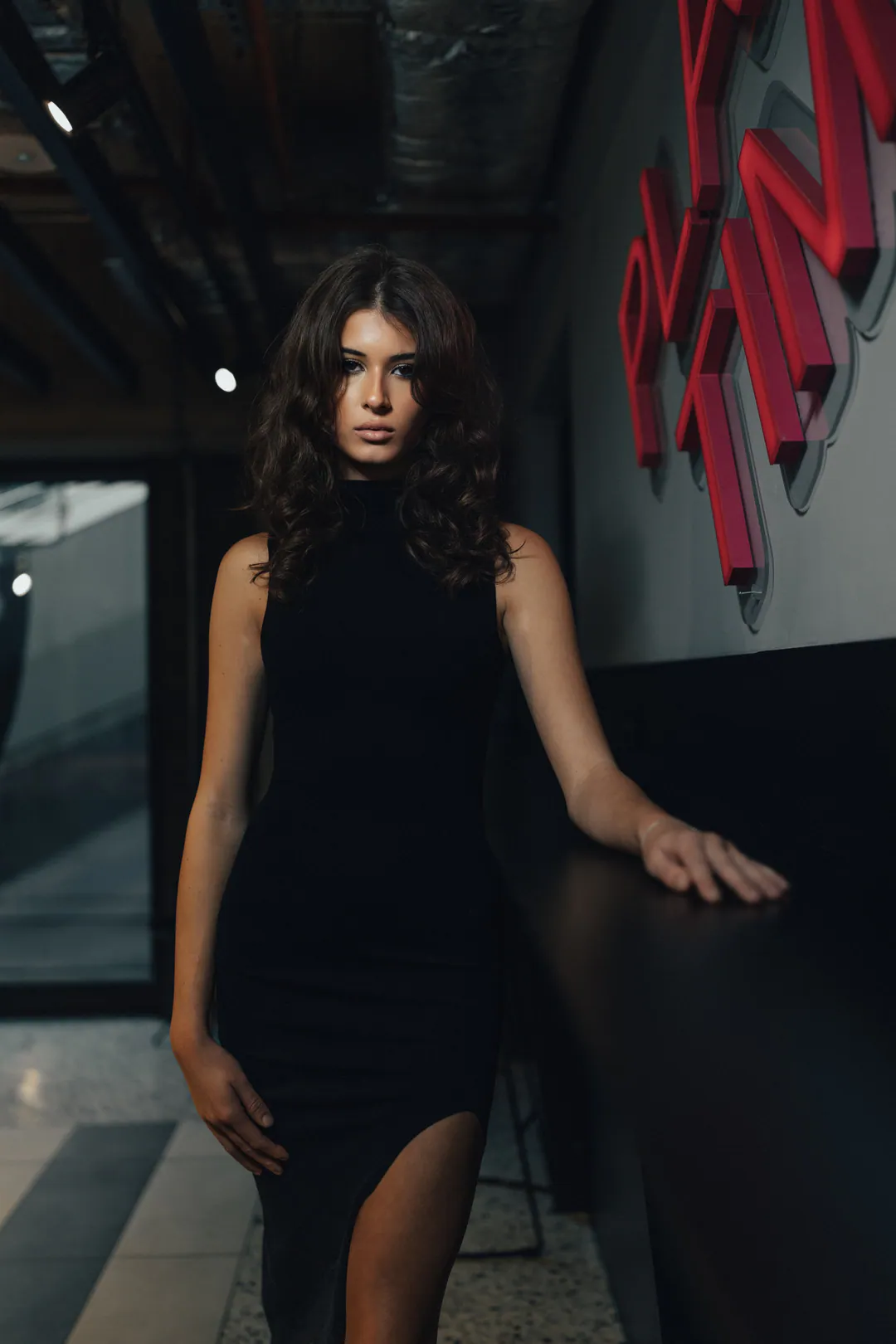 Woman in a black sleeveless split dress leaning on a bar beside red neon lettering in low light
