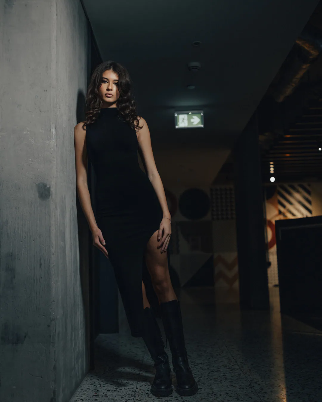 Woman in a black split dress and knee-high boots leaning against a concrete pillar in a dark corridor